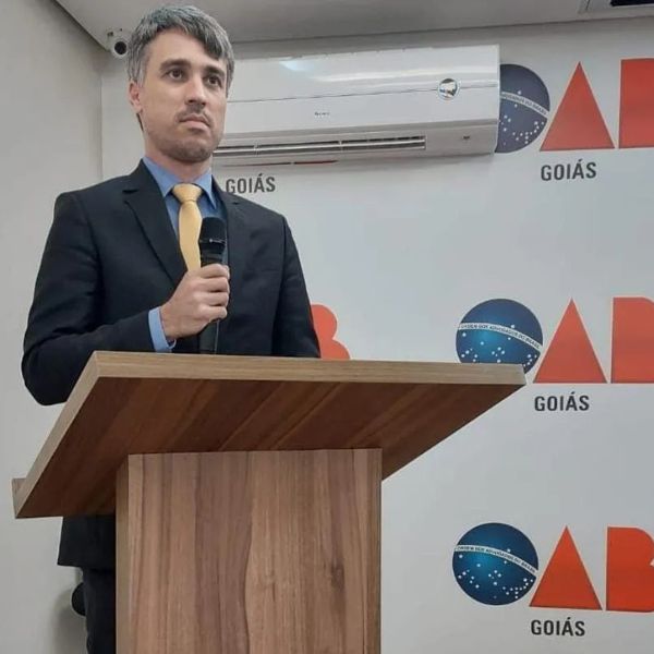 Retrato profissional de Pedro Rafael de Moura Meireles, advogado trabalhista, homem de meia-idade em traje social formal, terno escuro e gravata amarela, discursando ao microfone em um púlpito de madeira, fundo com logo institucional da OAB Goiás, ambiente sério e elegante, fotografia realista